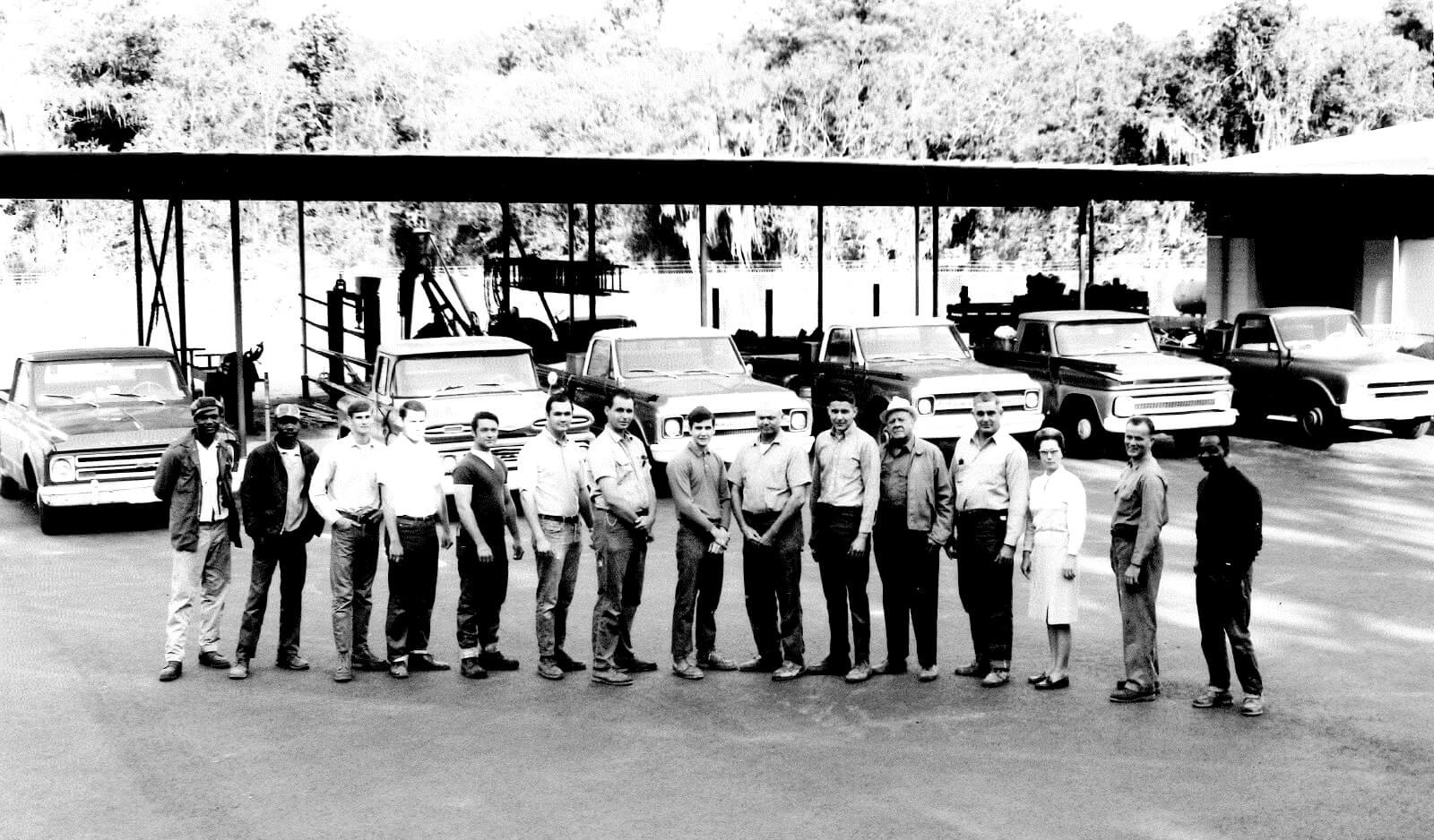 Black-and-white photograph of thirteen Hutson Plumbing crew members standing in a line in front of the pipe rack and truck fleet, April 1969