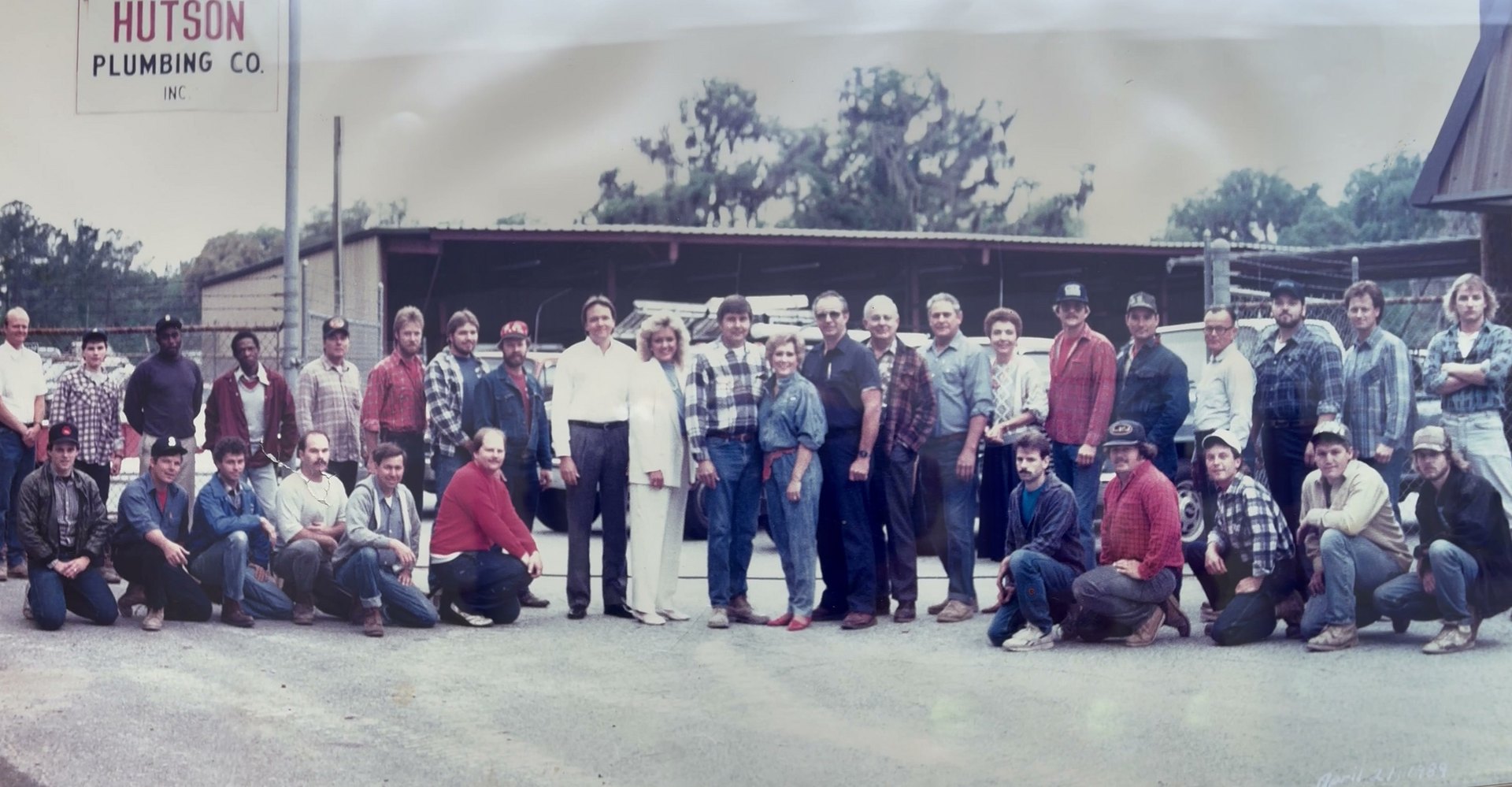 Color photograph of the whole Hutson Plumbing crew, thirty-three people in two rows, in front of the shop and the company sign, April 1989