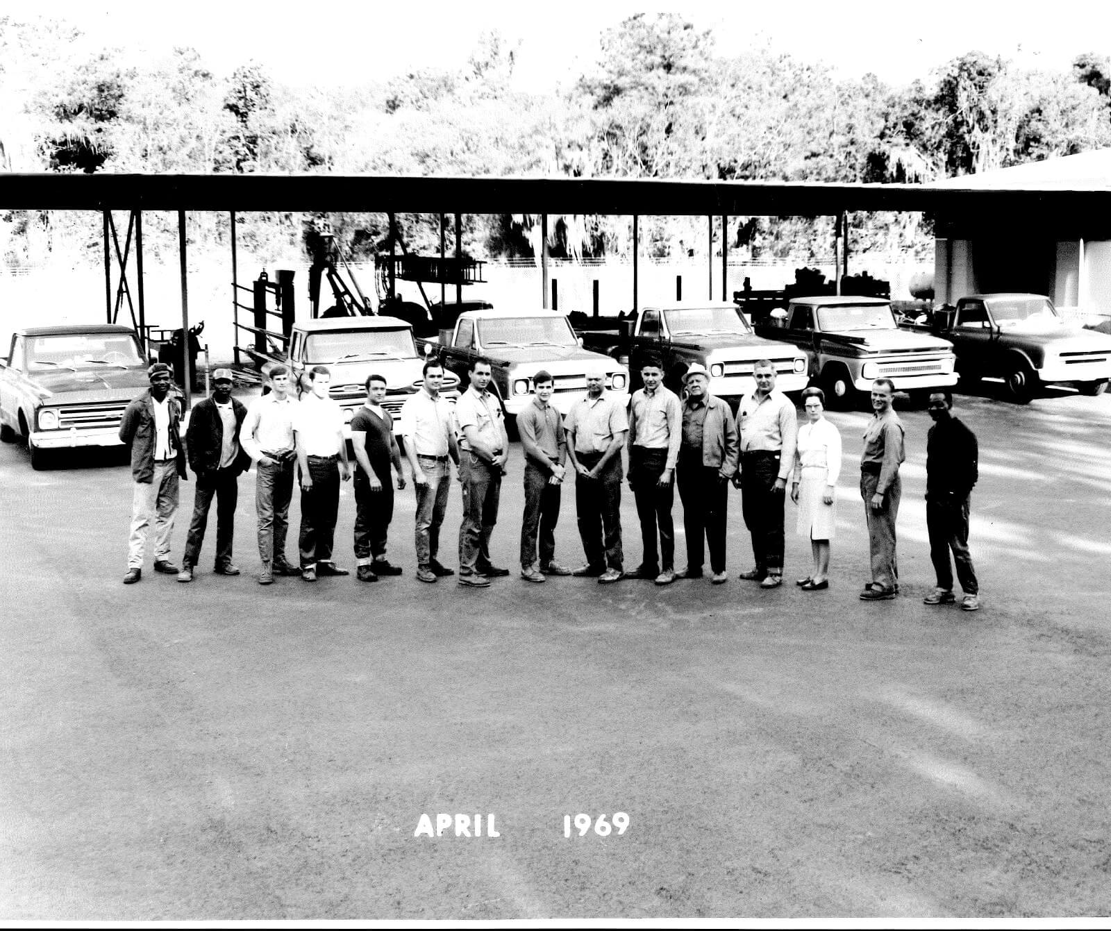 Black-and-white photograph of thirteen Hutson Plumbing crew members standing in a line in front of the pipe rack and truck fleet, captioned April 1969